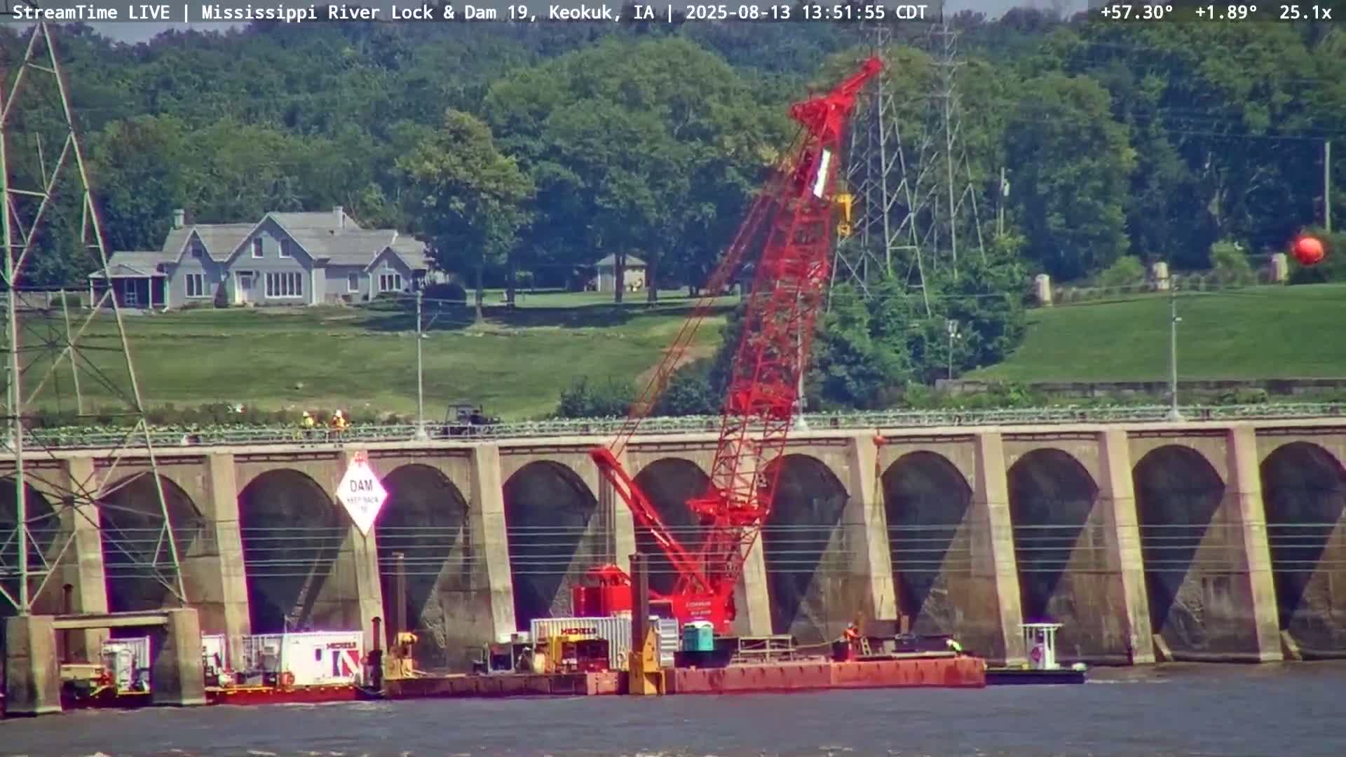 A red crane on a barge is positioned in front of a concrete dam on a sunny day.