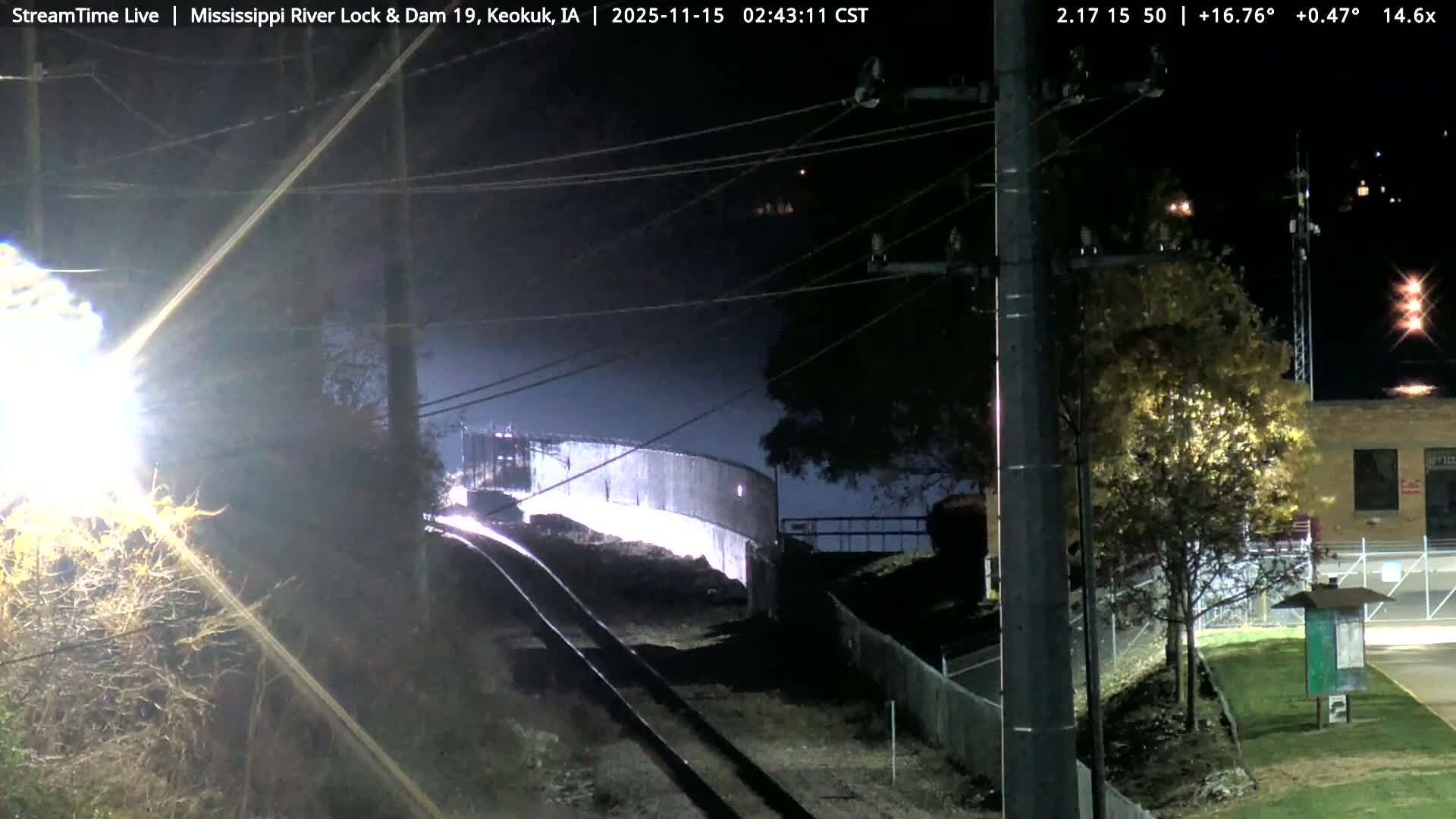 Under a clear night sky, railroad tracks lead towards a brightly lit, curved industrial structure, flanked by utility poles, a building, and trees, with a strong light source creating significant glare on the left.
