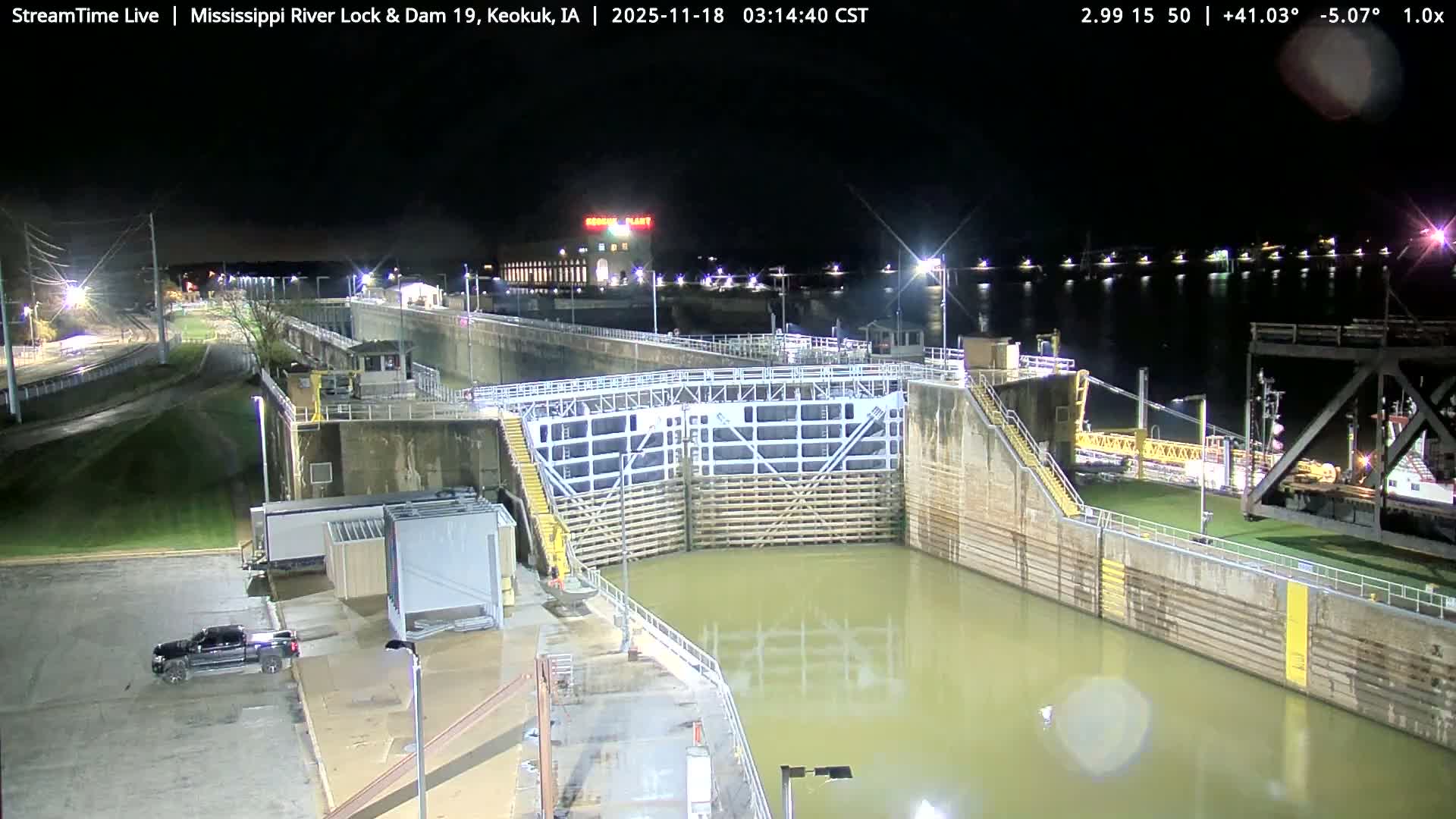 Under a clear night sky, railroad tracks lead towards a brightly lit, curved industrial structure, flanked by utility poles, a building, and trees, with a strong light source creating significant glare on the left.