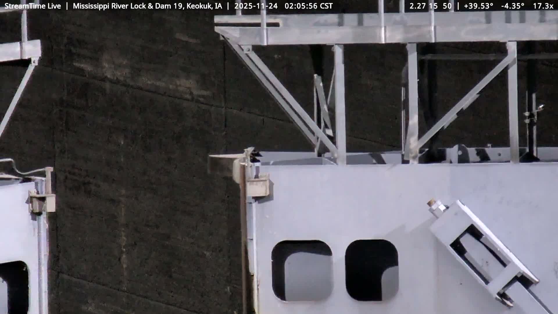A close-up view reveals a massive dark, textured concrete wall in the background, partially obscured by an intricate arrangement of light-colored metal structural beams, panels with rounded openings, and various mechanical components in the foreground, under overcast conditions.