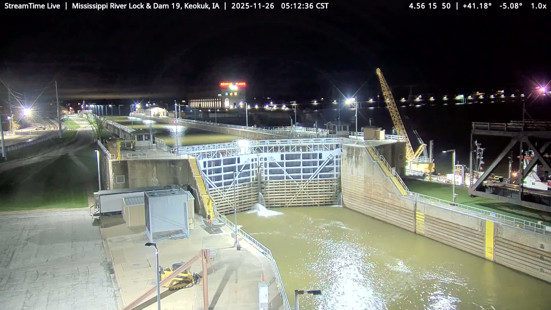 A close-up view reveals a massive dark, textured concrete wall in the background, partially obscured by an intricate arrangement of light-colored metal structural beams, panels with rounded openings, and various mechanical components in the foreground, under overcast conditions.