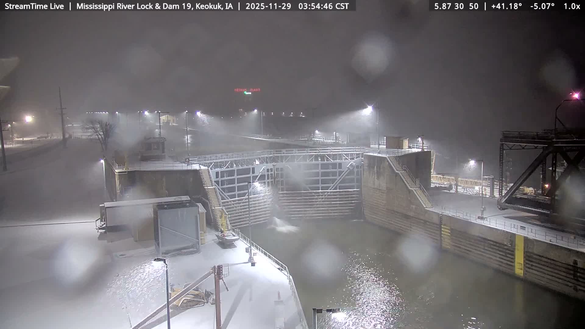 A river lock and dam system is illuminated by numerous artificial lights on a dark, snowy night, with accumulated snow covering the ground and structures, and visible precipitation in the air.