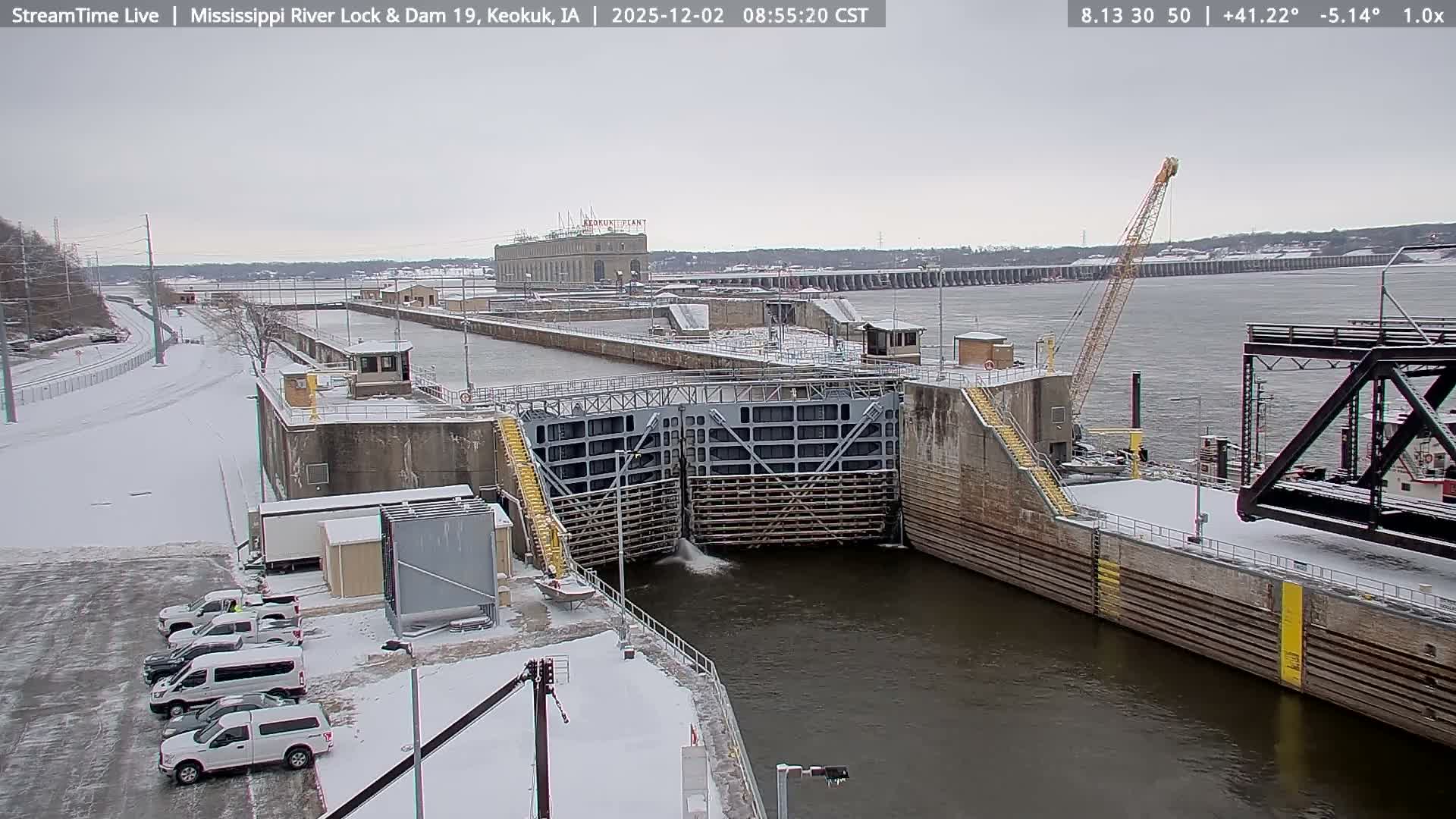 On an overcast winter day, a snow-covered river lock and dam complex shows water actively flowing through its partially open gates, with ice on the distant river and parked vehicles coated in snow.