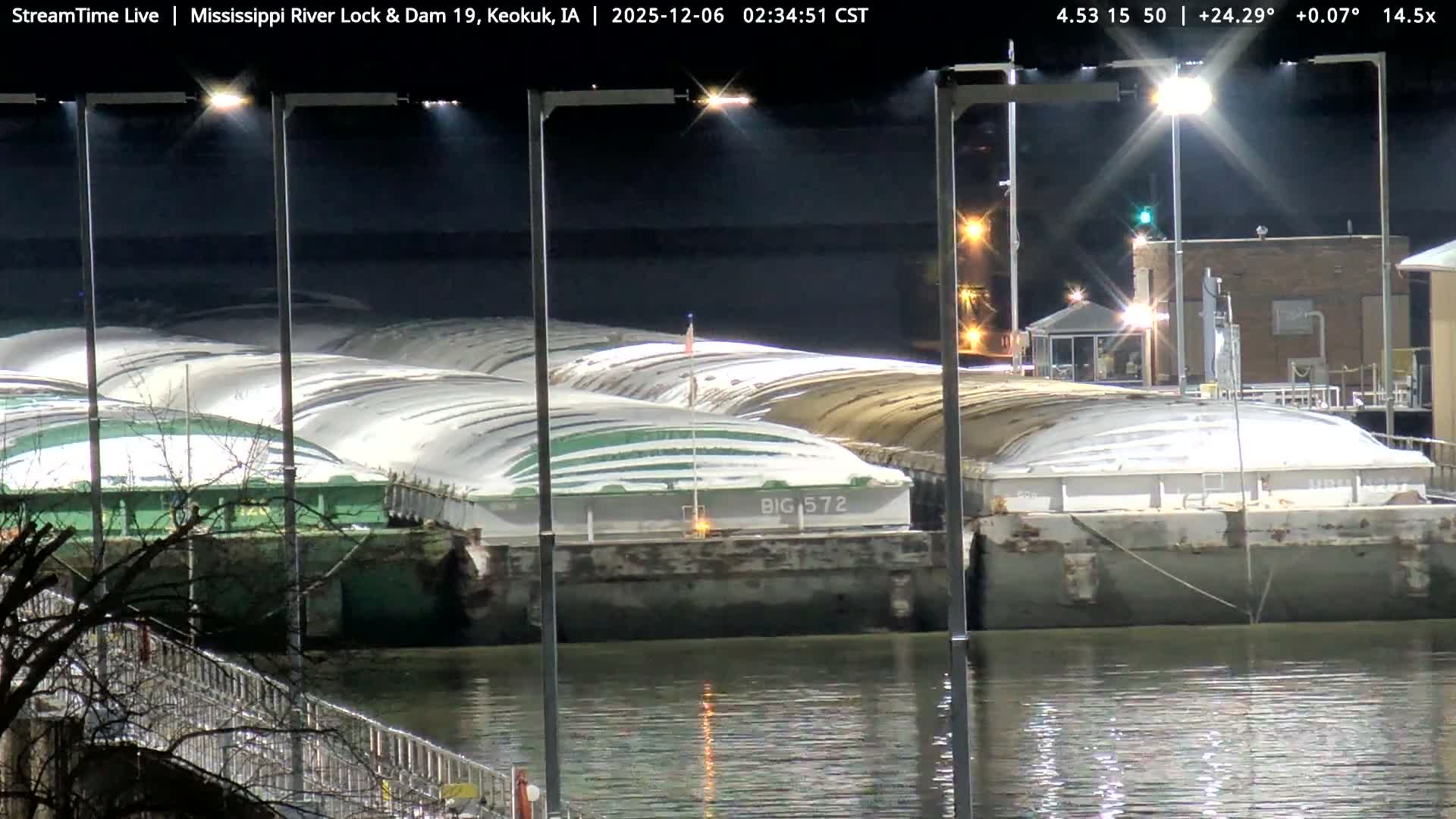 Numerous industrial barges, many covered with large white and green tarps, rest on a calm river at night, brightly illuminated by dock lights under a clear, dark sky.