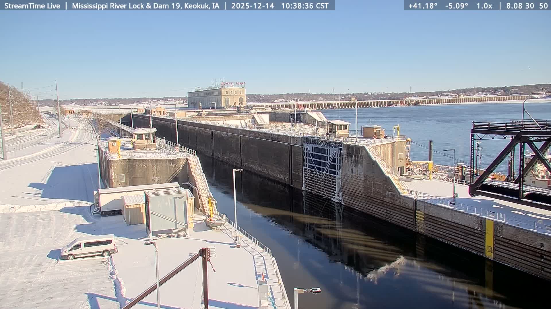 A brightly lit river lock and dam is seen at night with snow covering the ground, bare trees in the foreground, and turbulent water flowing over the dam under a clear, cold sky.