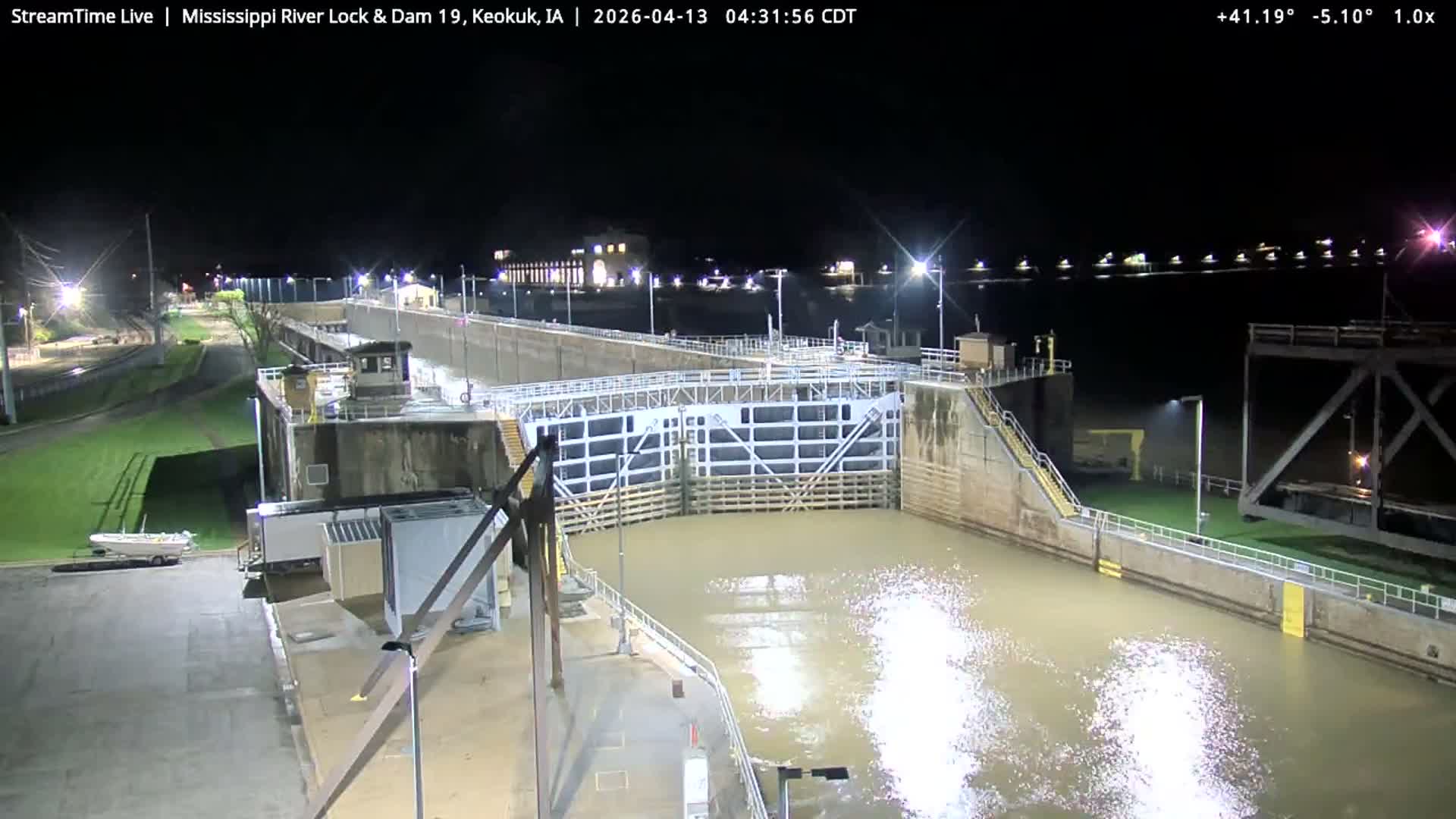 A brightly lit river lock and dam is seen at night with snow covering the ground, bare trees in the foreground, and turbulent water flowing over the dam under a clear, cold sky.