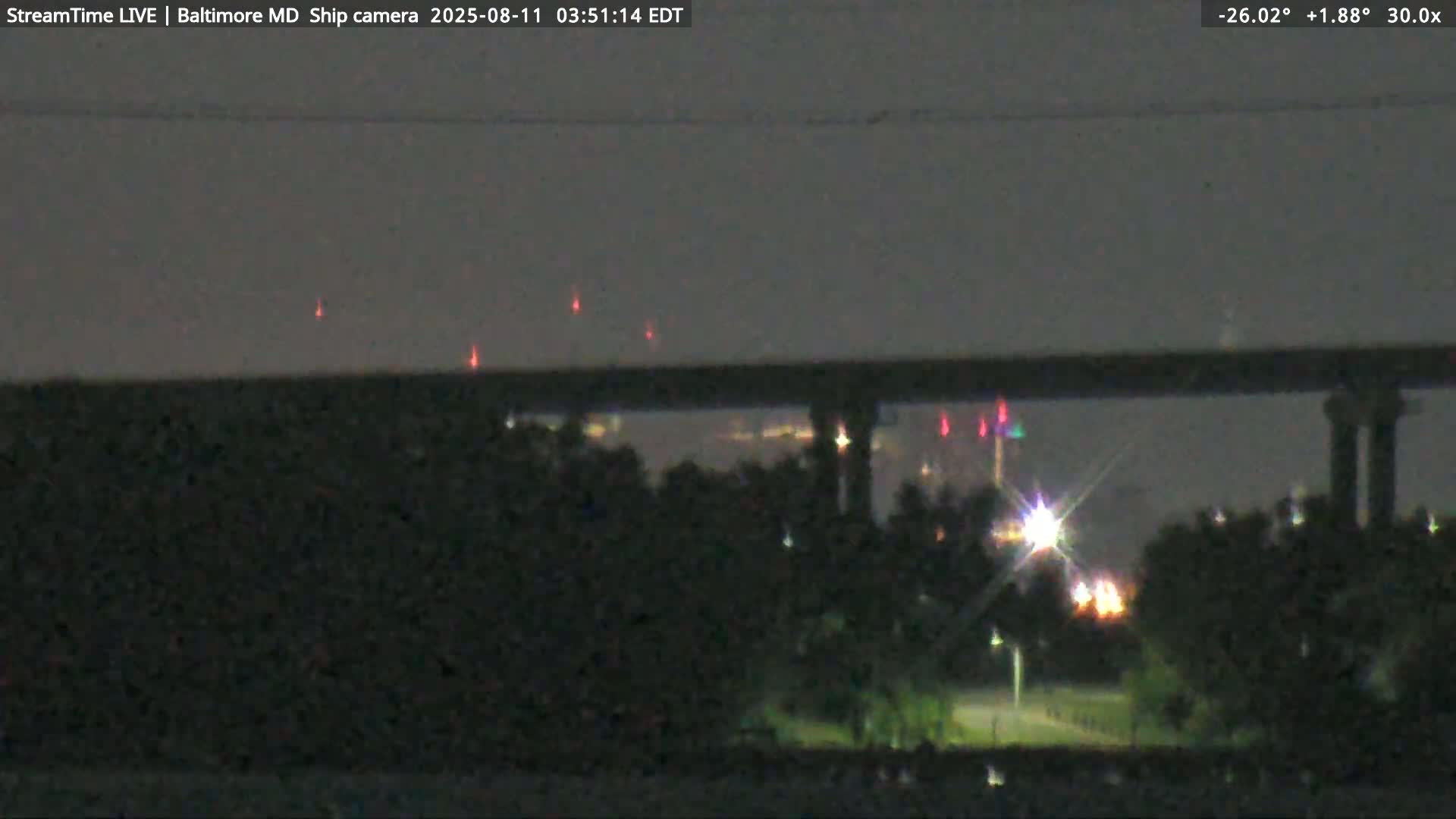 At night, a dark bridge is seen over a body of water with a few lights visible on the far shore and navigation lights on the bridge.