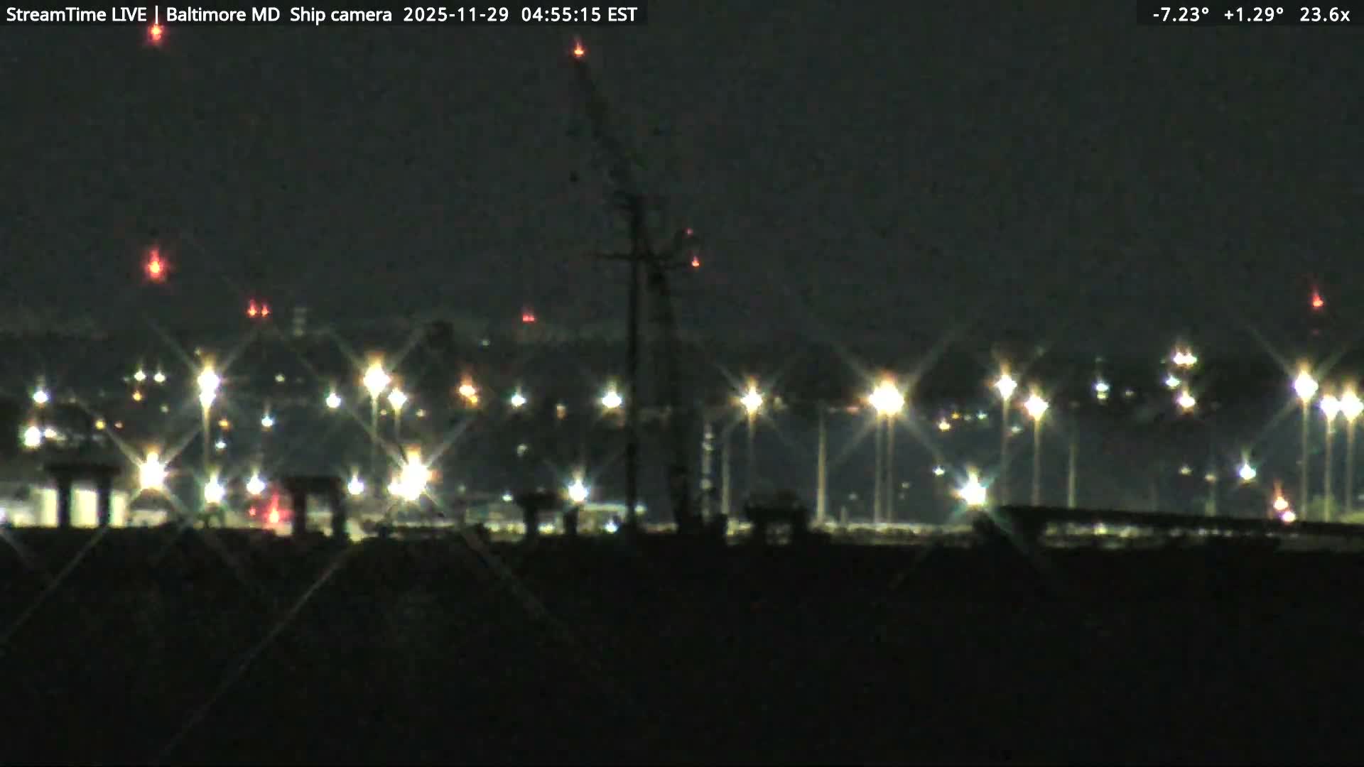 A clear nighttime view reveals a distant cityscape or industrial area brightly illuminated by numerous lights, with a tall dark tower silhouette topped by red lights standing in the midground, under a dark, clear sky.