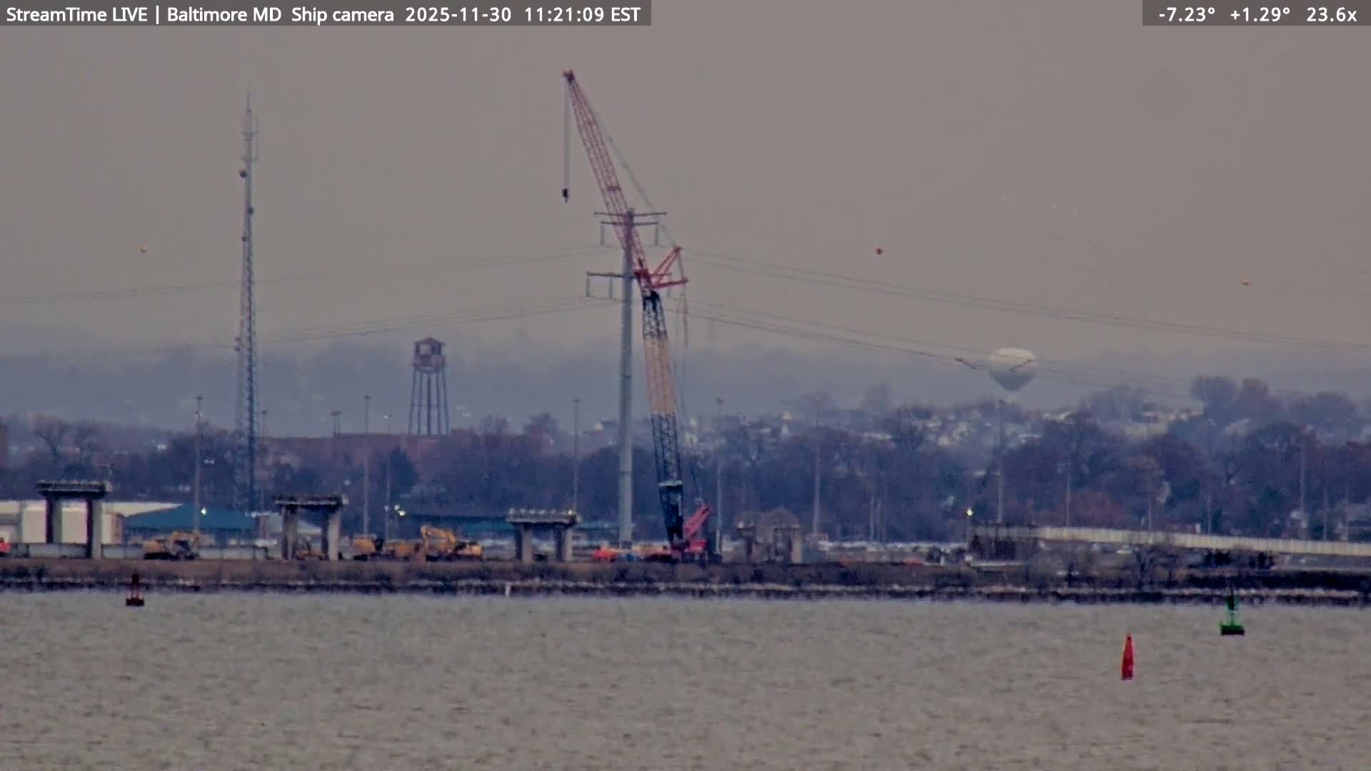 A clear nighttime view reveals a distant cityscape or industrial area brightly illuminated by numerous lights, with a tall dark tower silhouette topped by red lights standing in the midground, under a dark, clear sky.