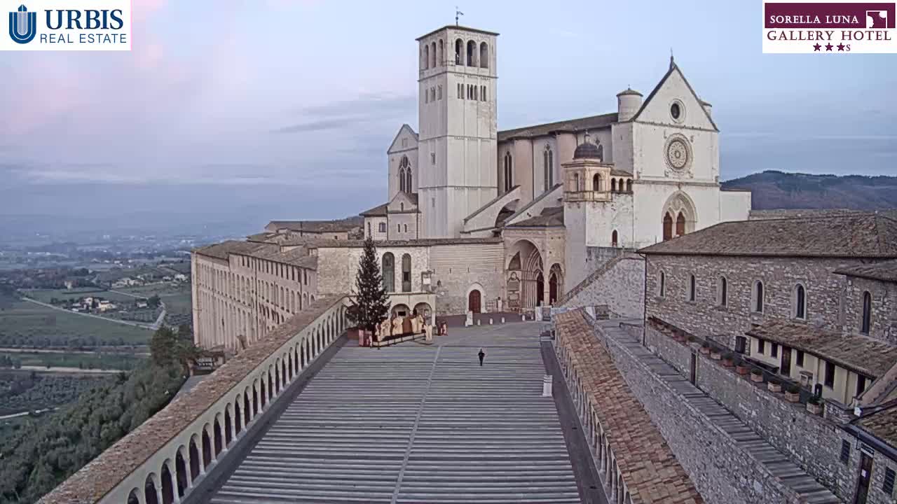 A large stone basilica sits atop a hill overlooking a valley under a hazy, pale sky, with a long staircase leading to its entrance.