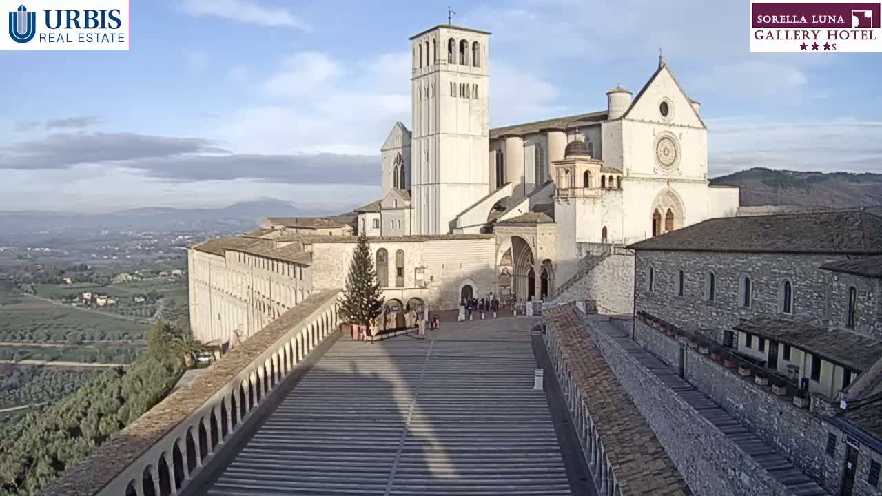 A large, light-colored basilica stands on a hill overlooking a valley under a mostly clear sky.