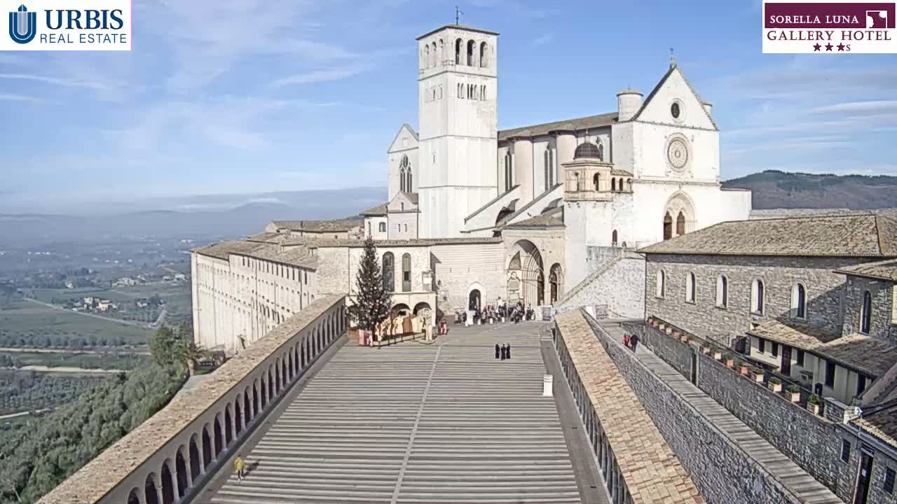 A large, light-colored basilica and surrounding buildings are viewed from above on a clear, sunny day, with a paved plaza in the foreground and a valley visible in the distance.