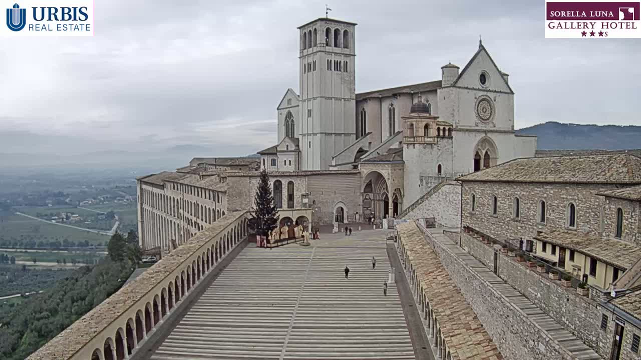 A large stone basilica and adjoining buildings sit atop a hill overlooking a valley on an overcast day.