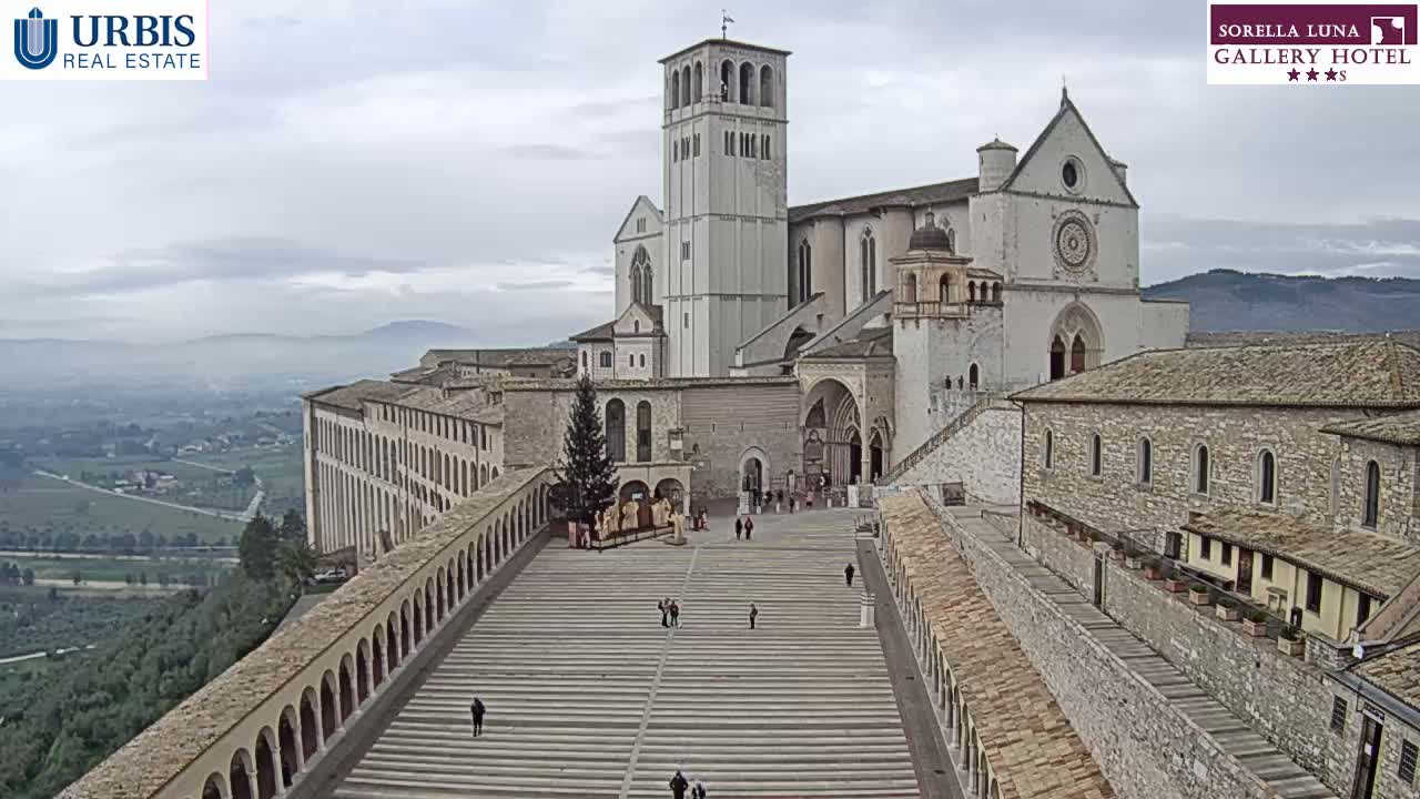 A large stone basilica and adjoining buildings are viewed from above on an overcast day, with a wide plaza and long, arched walkways leading to the main entrance.