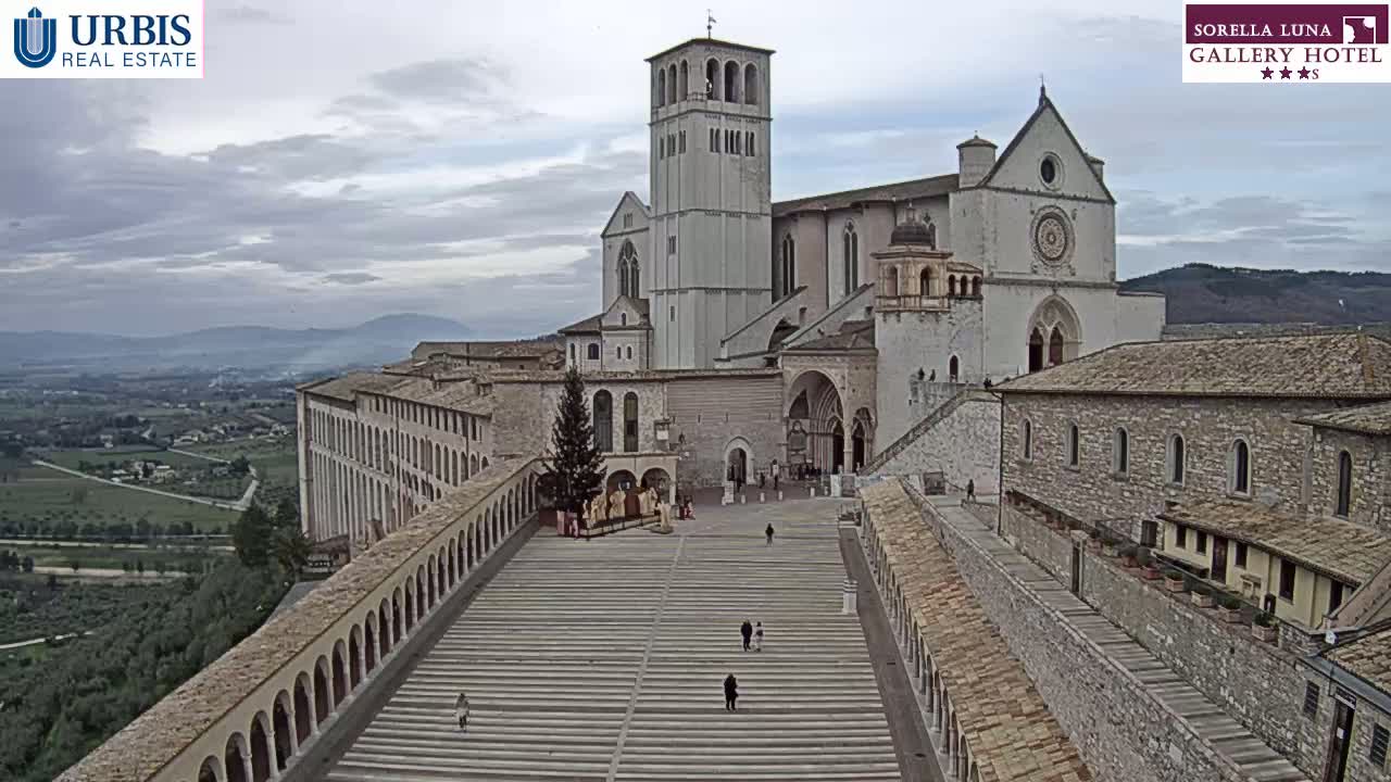 A large stone basilica and adjoining buildings sit on a hill overlooking a valley under an overcast sky.