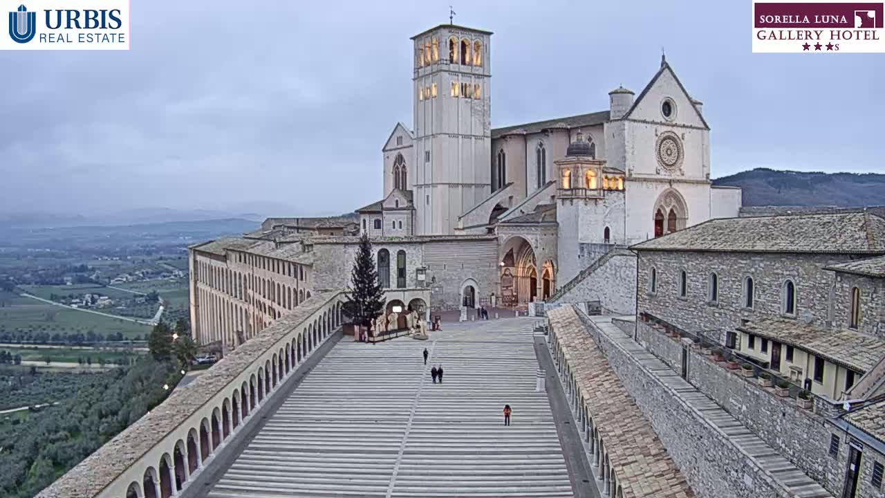 A large, light-colored stone basilica stands prominently on a hilltop overlooking a valley under an overcast sky, with a long, paved walkway leading down from its entrance.