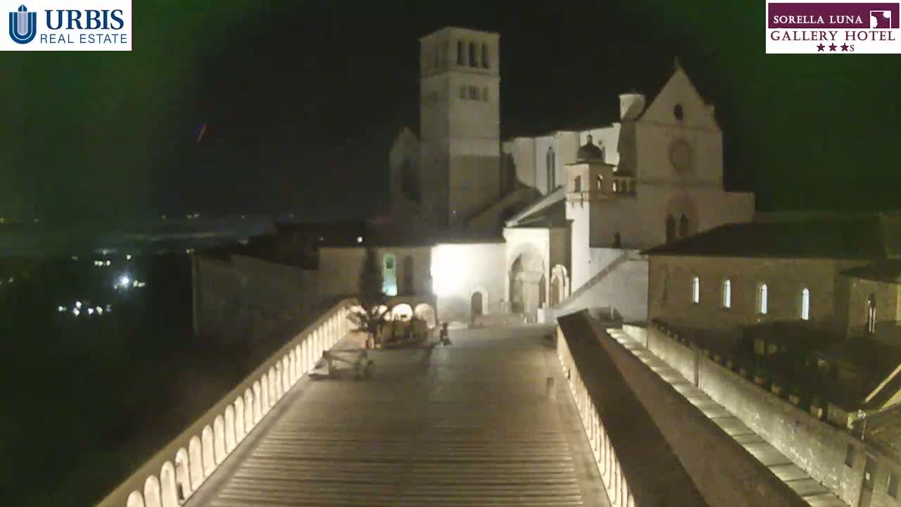 A large, illuminated stone building complex is viewed from above at night, with a long, descending pathway leading towards it.