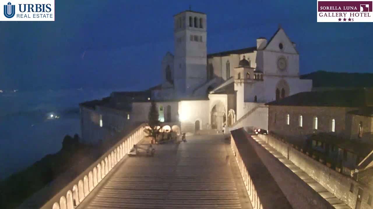 A large, illuminated stone basilica sits atop a hill at night, overlooking a valley shrouded in fog.
