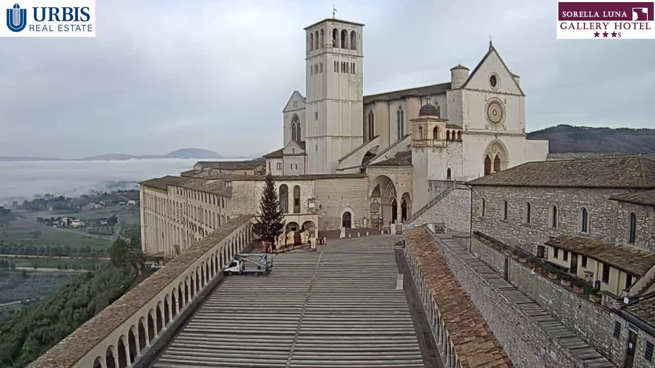 A large stone basilica sits atop a hill overlooking a valley shrouded in fog under an overcast sky.