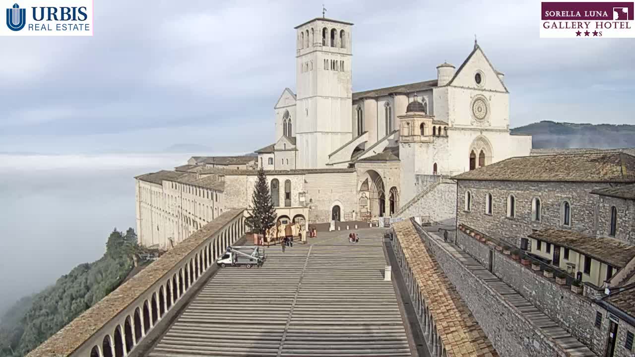 A large, light-colored stone basilica sits atop a hill overlooking a low-lying fog, with other stone buildings and a wide plaza in the foreground.