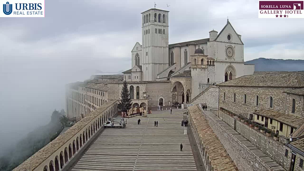 A large stone basilica stands in a town square, partially obscured by fog rolling in from the valley below.