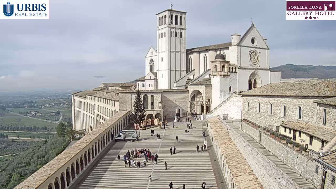 A large, light-colored basilica and adjoining buildings overlook a paved plaza with a few people, under a mostly clear sky.