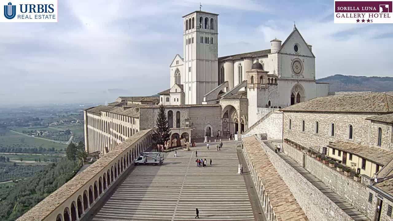 A large stone basilica and adjoining buildings stand on a hill overlooking a valley on a partly cloudy day.