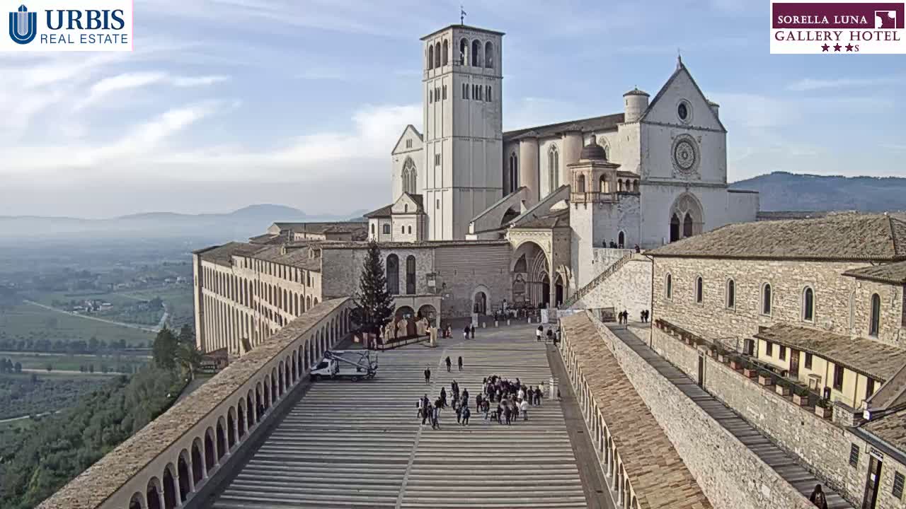 A large stone basilica with a tall bell tower is seen from above on a partly cloudy day, with a spacious plaza in front and a wide view of a valley in the background.