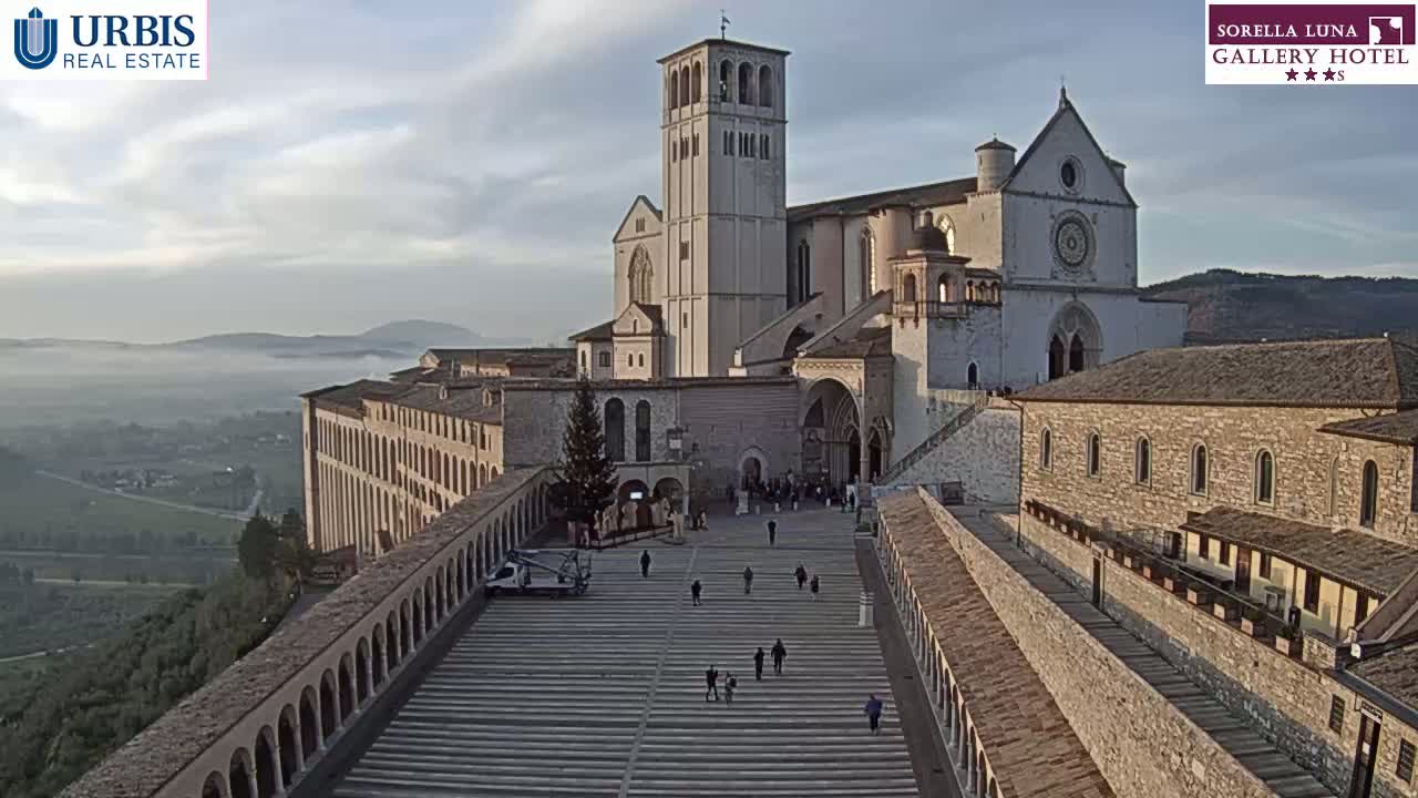 A large stone basilica and surrounding buildings are situated on a hill overlooking a fog-shrouded valley under a partly cloudy sky.