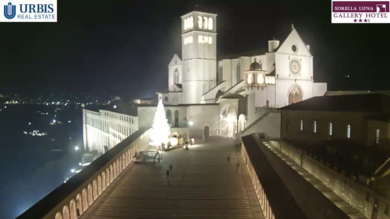 A large, illuminated basilica and a Christmas tree are visible at night from a high vantage point overlooking a town.