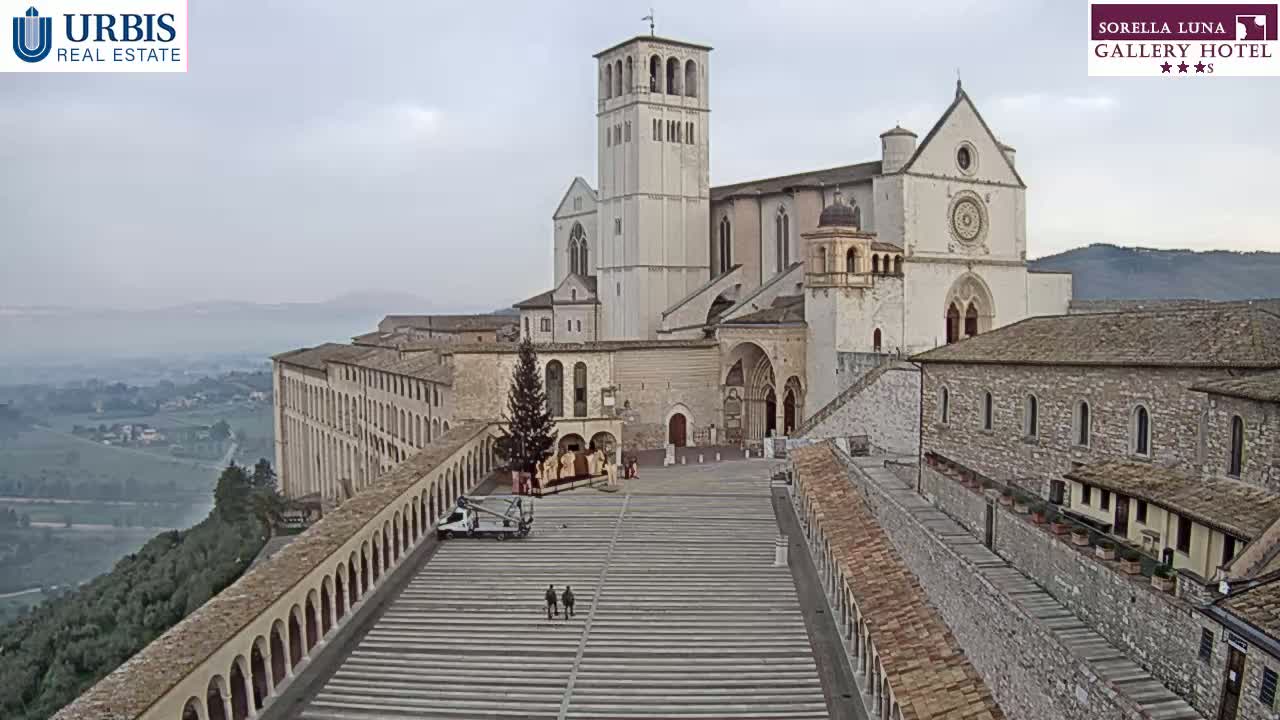 A large stone basilica and adjoining buildings sit atop a hill overlooking a valley on a hazy, overcast day.
