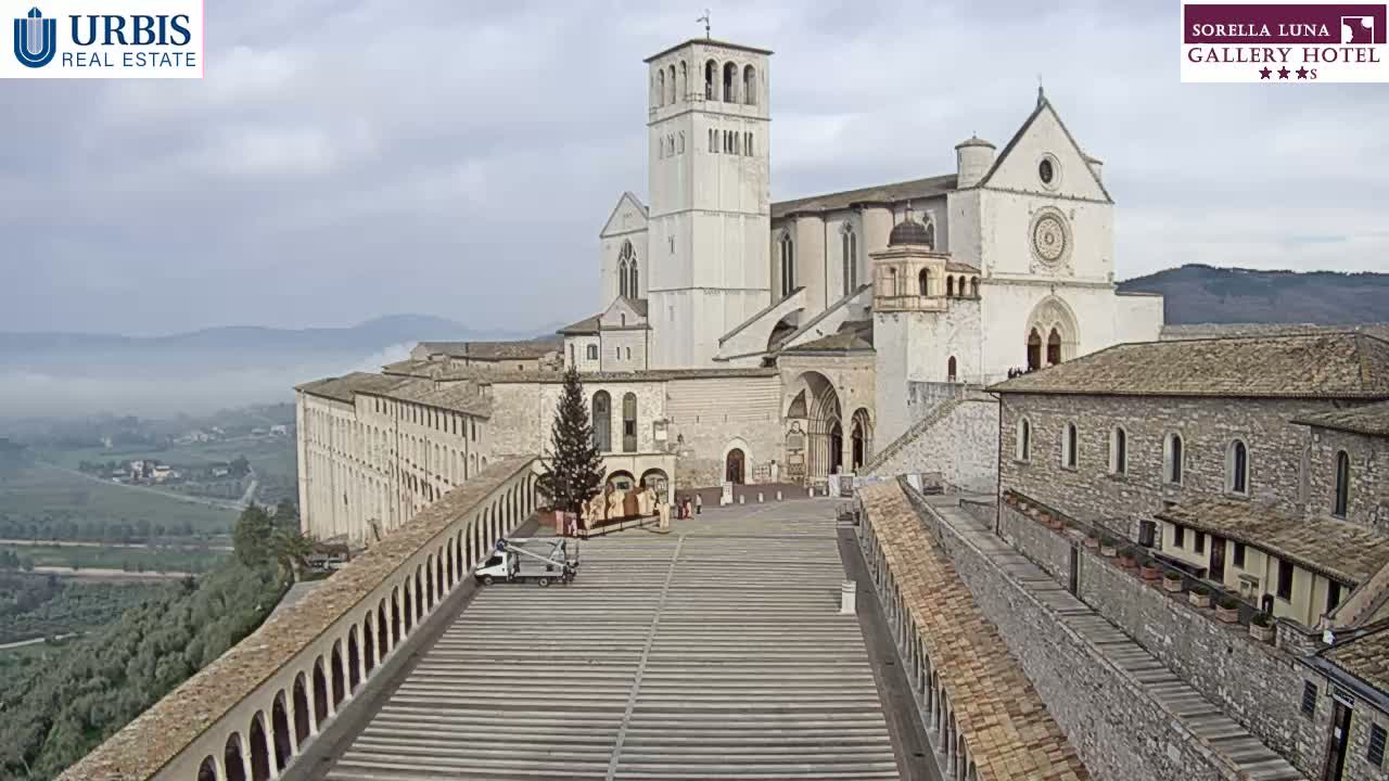 A large, light-colored basilica stands prominently in a town overlooking a valley shrouded in fog, under a cloudy sky.