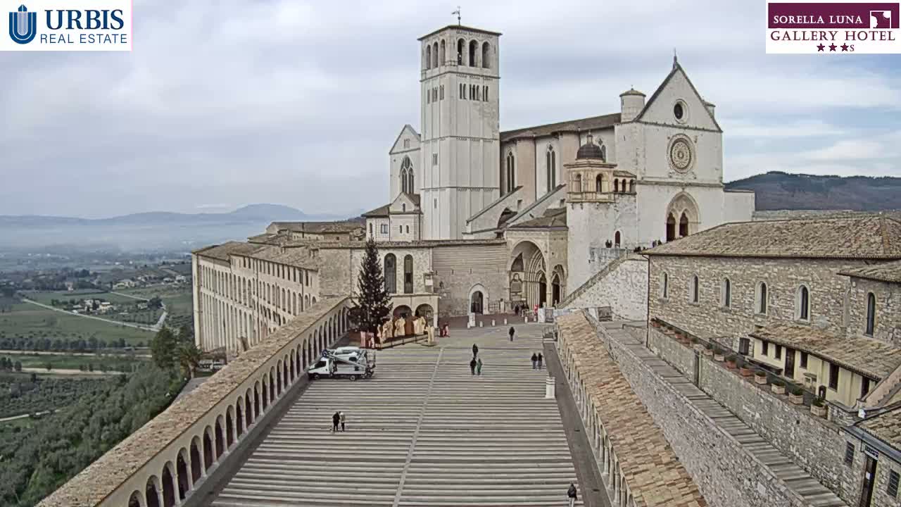 A large stone basilica and adjacent buildings overlook a wide plaza and a valley on an overcast day.