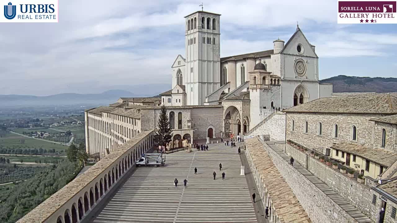 A large, light-colored basilica and adjoining buildings are viewed from above on a partly cloudy day, with a plaza and surrounding landscape visible in the distance.
