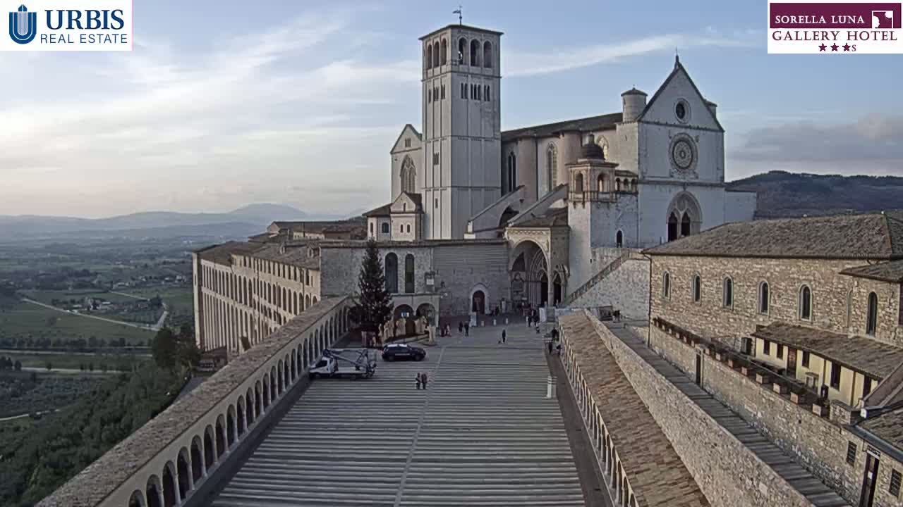 A large stone basilica sits atop a hill overlooking a valley under a partly cloudy sky, with a paved plaza in front of it and a few people walking around.