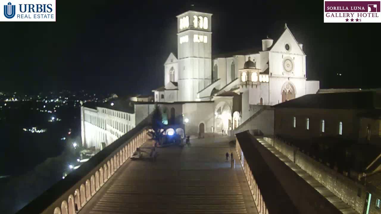 A large, illuminated basilica is seen at night from an elevated viewpoint, with a paved walkway leading down to it.