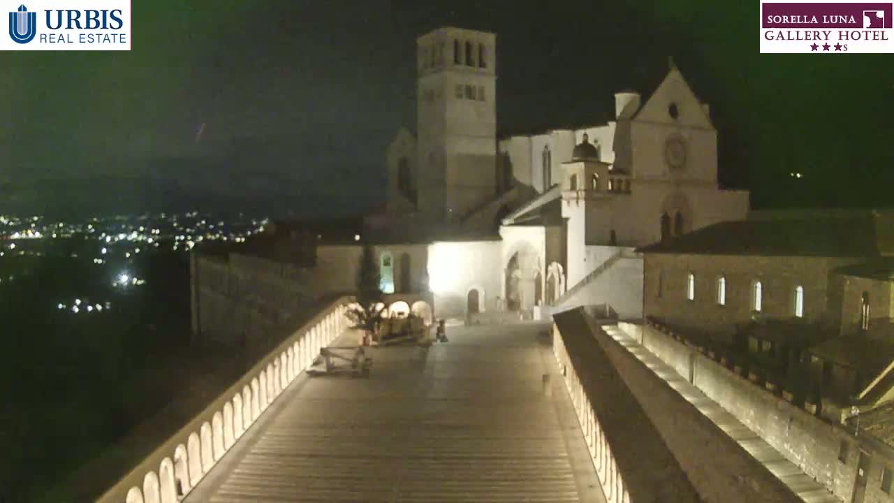 A large, illuminated basilica sits atop a hill at night, overlooking a city with a long, descending pathway lined with low walls.