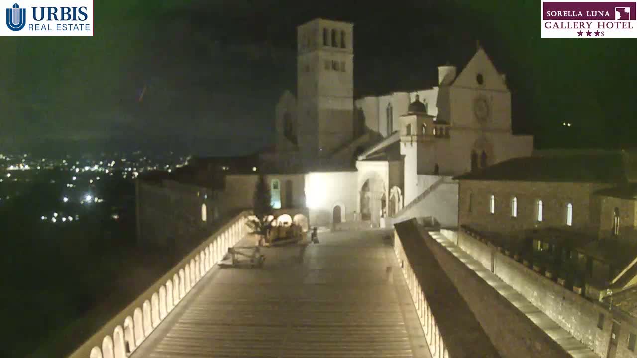 A large, illuminated building complex, possibly a church, stands at night, overlooking a city with lights visible in the distance under a clear, dark sky.