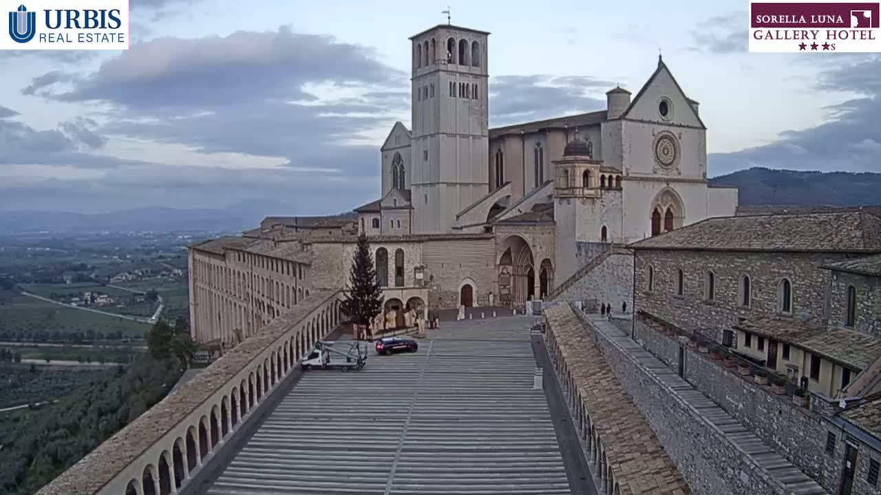 A large stone basilica sits atop a hill overlooking a valley under a cloudy sky, with a wide paved plaza in the foreground and a small Christmas tree visible near a parked car.