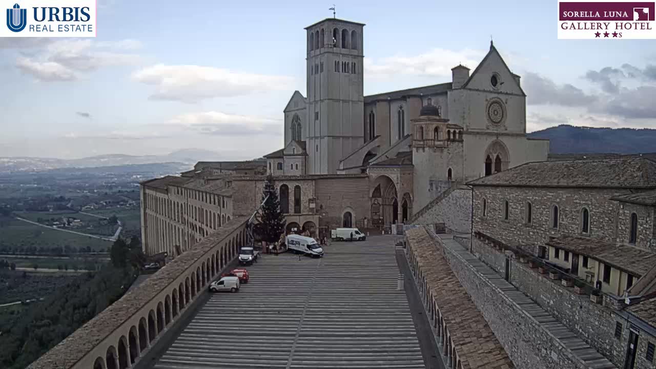 A large stone basilica and adjacent buildings are viewed from above on a partly cloudy day, with several vehicles parked in a plaza below.