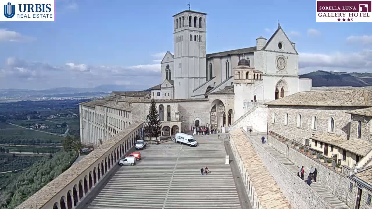 A large, light-colored basilica sits atop a hill overlooking a valley on a partly cloudy day, with a wide plaza in front of it and several vehicles parked nearby.