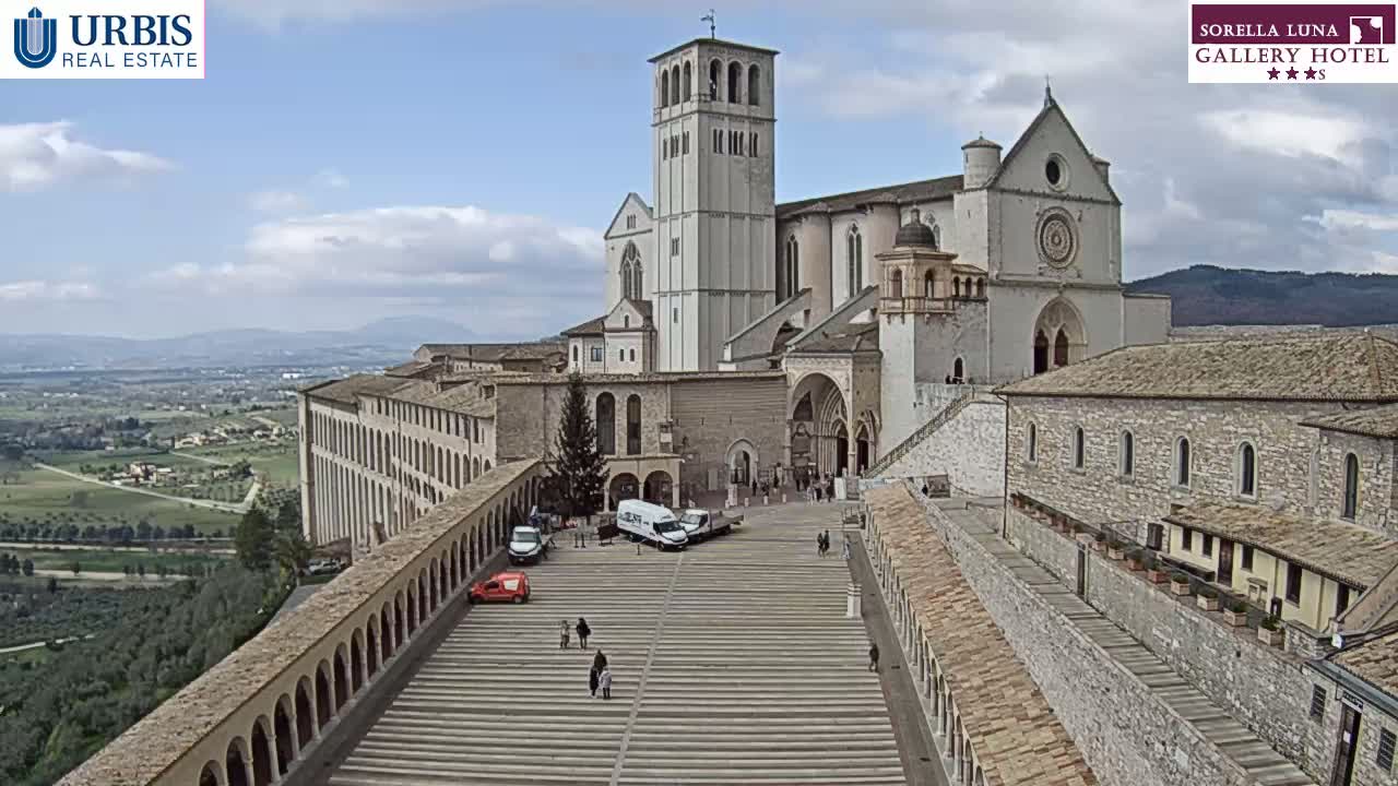 A large stone basilica with attached buildings and a plaza sits on a hill overlooking a valley under a partly cloudy sky.