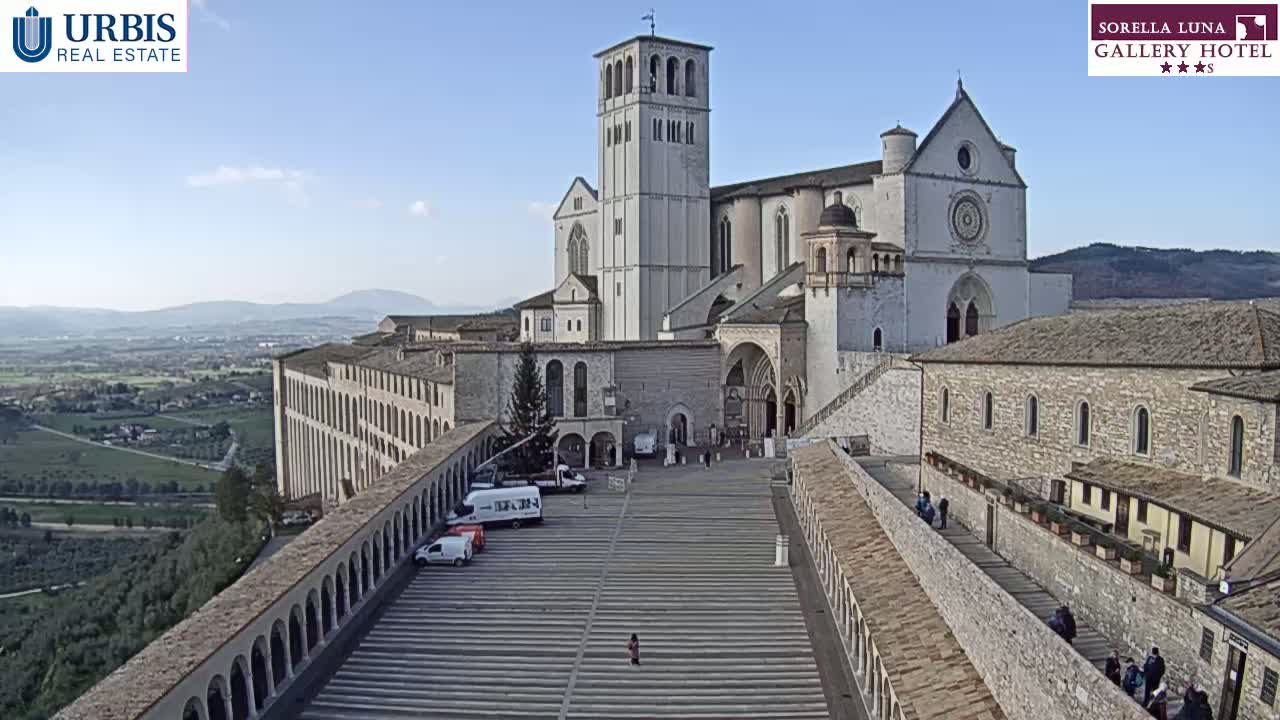 A large stone basilica sits atop a hill overlooking a valley under a clear blue sky, with a paved plaza in the foreground and several vehicles parked nearby.