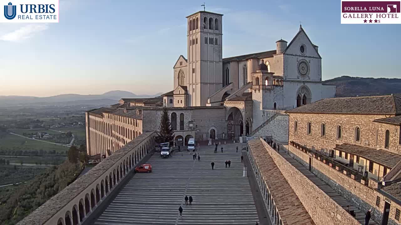 A large stone basilica and adjoining buildings are seen from above on a clear, sunny day, with a wide plaza and surrounding hills in the background.