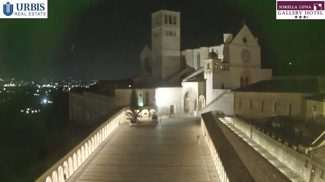 A nighttime view of a large stone building, possibly a church, illuminated against a dark sky with a distant city visible in the background.