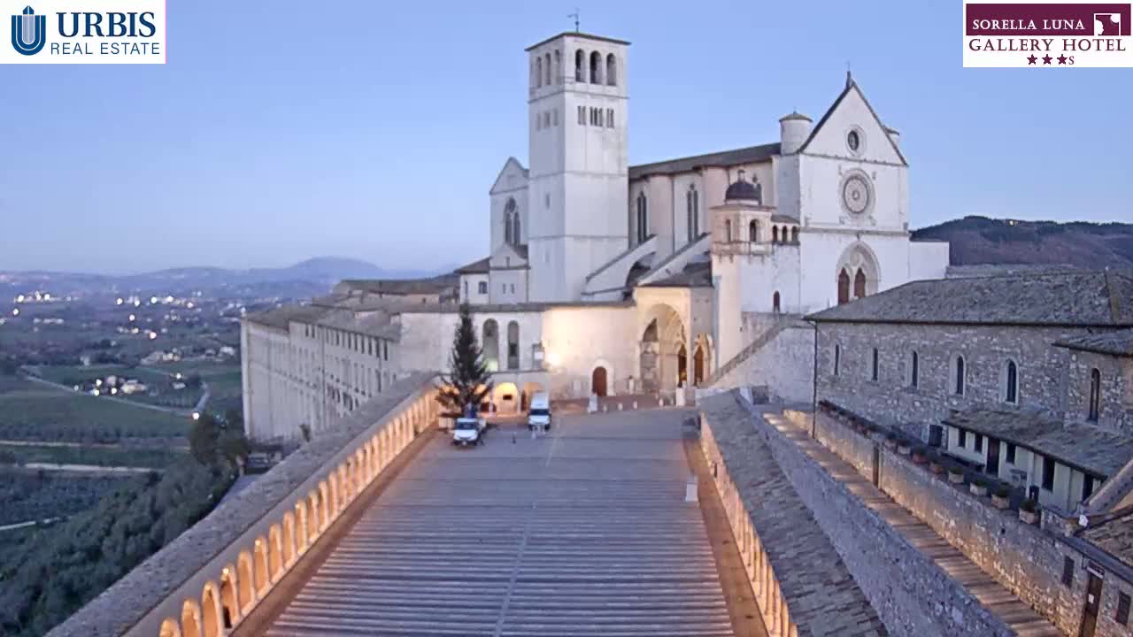 A large, light-colored basilica and adjacent buildings are situated on a hill overlooking a town at dusk, with a paved walkway leading down towards the city.