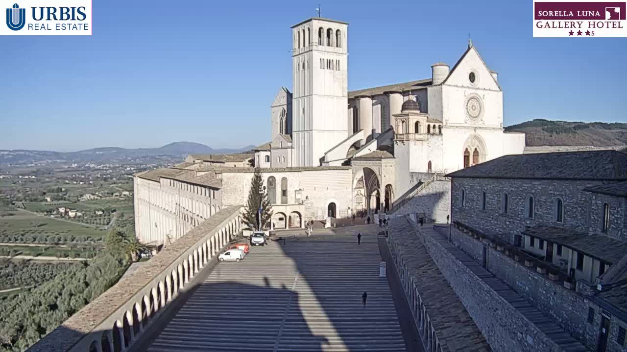 A large, light-colored basilica stands prominently in a sunlit cityscape, overlooking a valley under a clear blue sky.