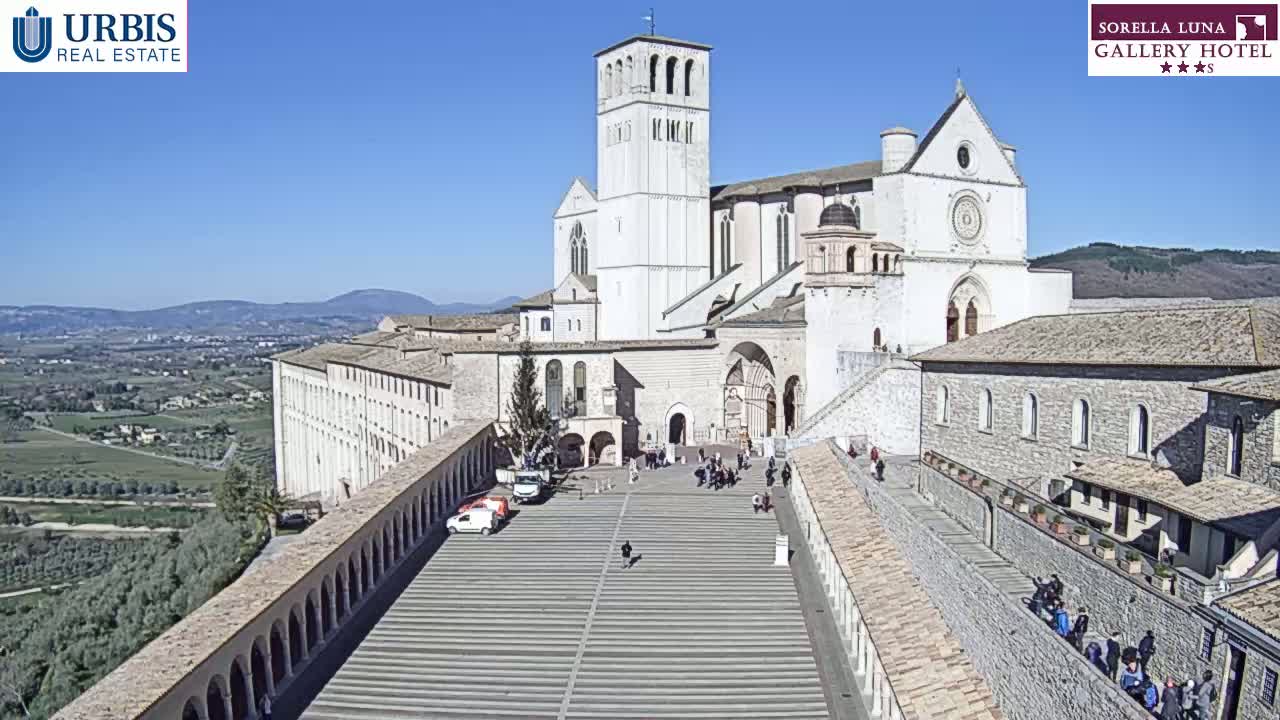 A large, light-colored basilica with a tall bell tower sits atop a hill overlooking a valley on a sunny day.