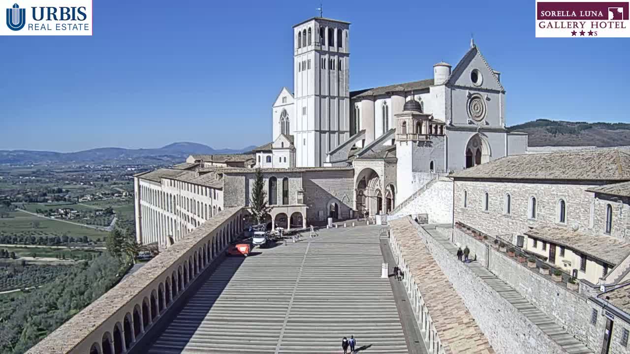 A large, light-colored stone basilica and adjacent buildings overlook a paved plaza and a valley under a clear blue sky.