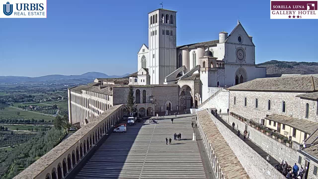 A large stone basilica and surrounding buildings are seen on a sunny day, overlooking a valley.